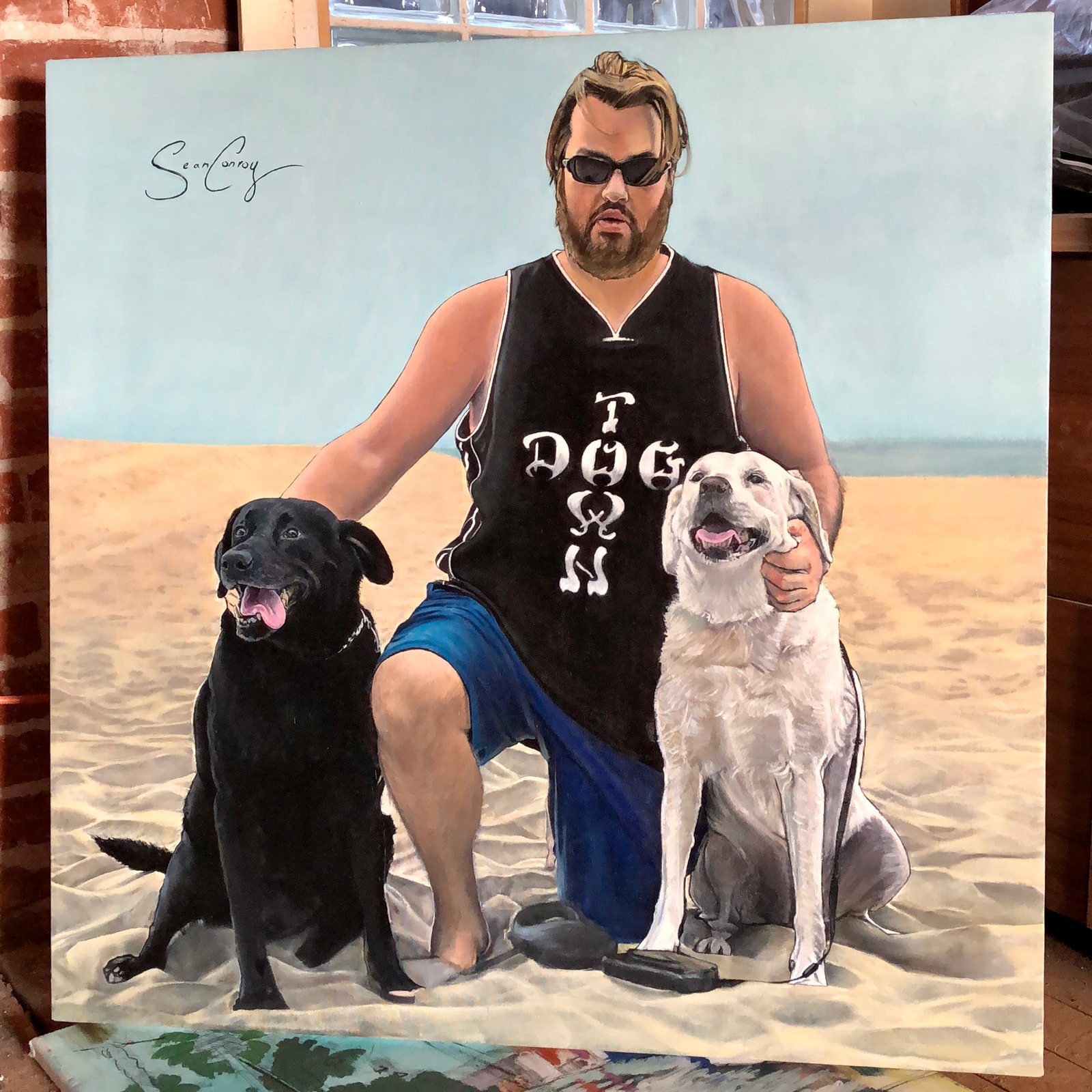 Portrait painting of a man seated on a beach with a black dog and a white dog.