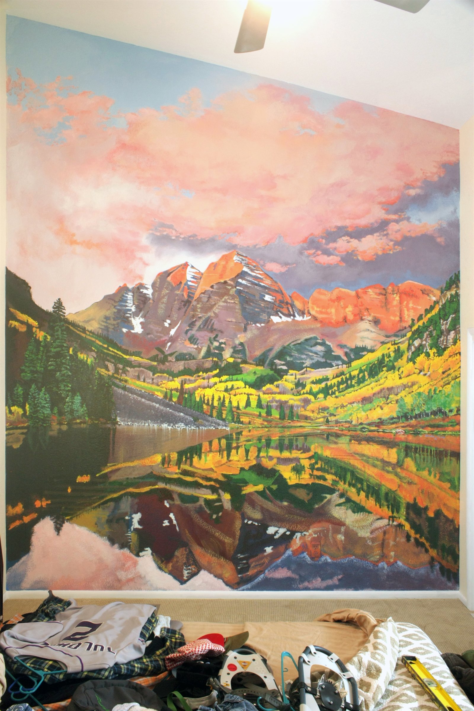 Large wall painting of mountain peaks, clouds, forest, lake reflections, and warm sunset colors.
